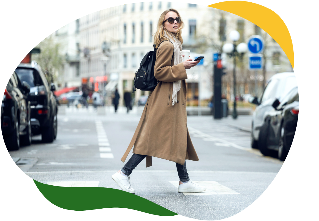 Suurte päikeseprillide ja beeži pika mantliga tänapäevane naine ületab tänavat, hoides paremas käes mobiiltelefoni.