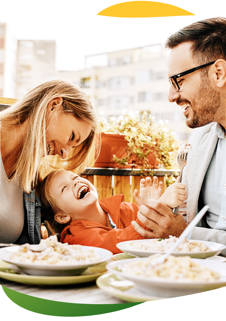 Tööriietes noored vanemad söövad söögilaua ääres, nende vahel istub laps. Nad naeravad ja neil on koos tore.